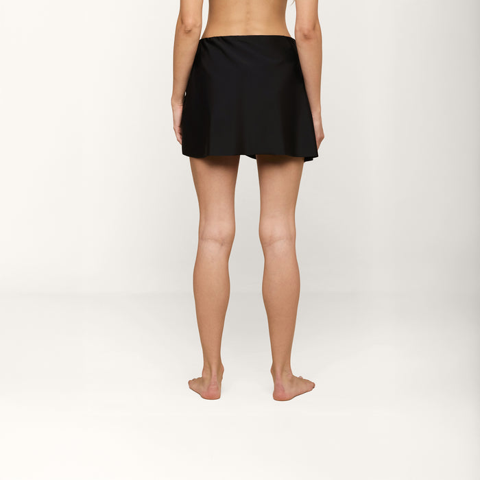 A person stands barefoot on a white surface, facing away from the camera, wearing the Chandra Cover-Up Skirt featuring gold hardware. The background is plain and light-colored.