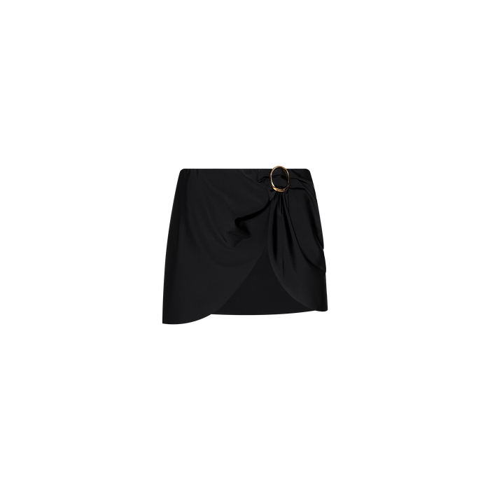 The Chandra Cover-Up Skirt is a black mini with an asymmetrical hem and side draping, accented by a decorative gold hardware buckle on the waistband. Displayed against a plain white background.