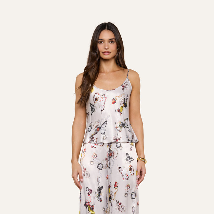 A woman wearing the Charmy Satin Camisole, a sleeveless pajama set featuring a white base with colorful floral and butterfly prints alongside abstract designs, stands against a plain background.