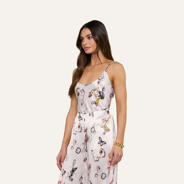 A woman with long brown hair models the Charmy Satin Camisole, a white satin jumpsuit adorned with vibrant butterfly prints and floral designs. The outfit features thin camisole straps and wide-leg pants. She is posed against a plain light background, facing slightly to the side.