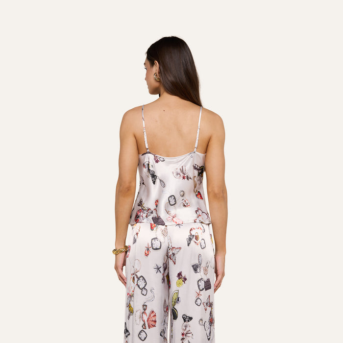 A woman with long brown hair, seen from behind, wears the Charmy Satin Camisole—a light-colored pajama top featuring a butterfly and floral print. She stands against a plain, light background.