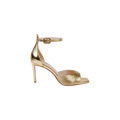 The Cici Stiletto Sandal features a gold high heel with an ankle strap and buckle, a square toe, and a textured band with leather pleating across the front. Finished in shiny metallic gold, its slender stiletto heel completes this elegant look.