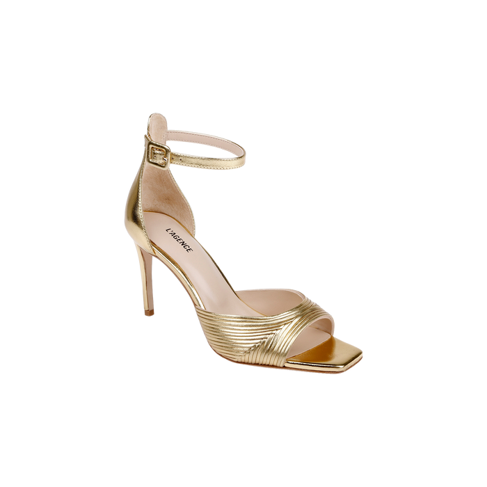 The Cici Stiletto Sandal is a gold high-heeled shoe featuring an ankle strap with a buckle, a square toe, and textured crisscross straps over the front.