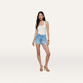 A woman with long dark hair and a neutral expression stands against a plain background wearing a white tank top, gold heeled sandals, gold bracelets, and the Clark Distressed Jean Short in a light wash high rise style.