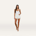 A woman stands facing forward against a plain white background, wearing a white tank top, nude heels, and the Clark Distressed Jean Short—an all-cotton, high-rise denim short—with one hand in her pocket.