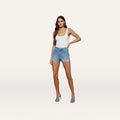 A woman wearing a white tank top, the Clark Distressed Jean Short—high-rise blue shorts made from all-cotton denim—and blue high-heeled sandals stands against a plain white background.