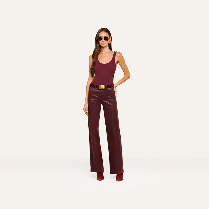 A woman wearing sunglasses, a sleeveless maroon top, the Clayton Coated Wide-Leg Jean with a contoured waistband, a belt with a gold buckle, and matching boots stands against an off-white background.