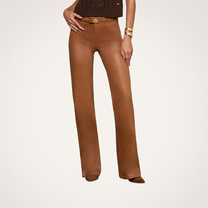 A person wearing the Clayton Coated Wide-Leg Jean, featuring a contoured waistband in brown leather, paired with a brown top, brown shoes, and gold bracelets stands against a plain white background. The upper part of the head is not visible.