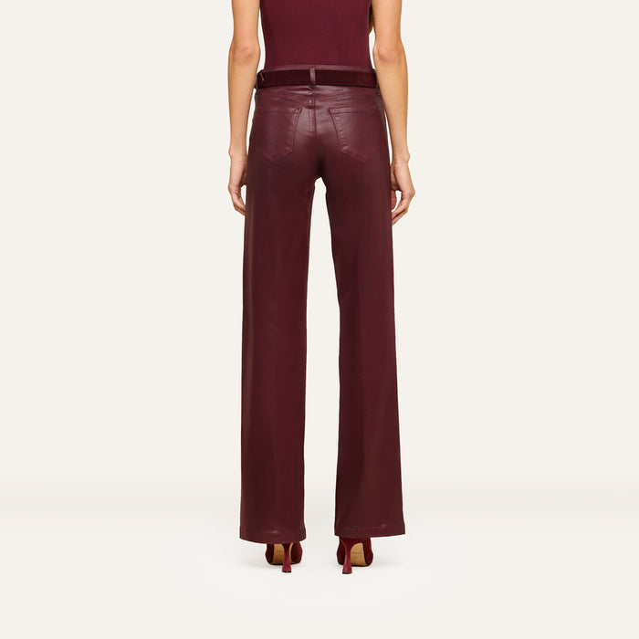 A person stands facing away, wearing the Clayton Coated Wide-Leg Jean featuring a contoured waistband in a rich burgundy hue, paired with a matching belt and heels. The background is plain and light-colored.