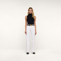 A woman stands facing forward against a plain white background, wearing a sleeveless black turtleneck, a black belt with a large gold buckle, black shoes, and the Clayton Wide-Leg Jean in white.
