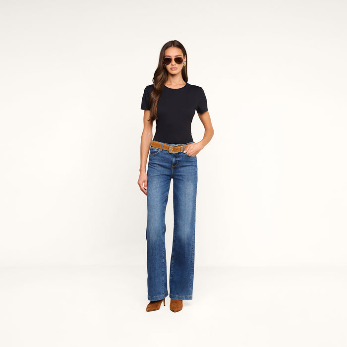 A woman with long hair stands against a plain white background, wearing sunglasses, a black short-sleeve shirt, brown heeled shoes, and the Clayton Wide-Leg Jean in a high-rise blue wash with a brown belt; one hand rests in her pocket.