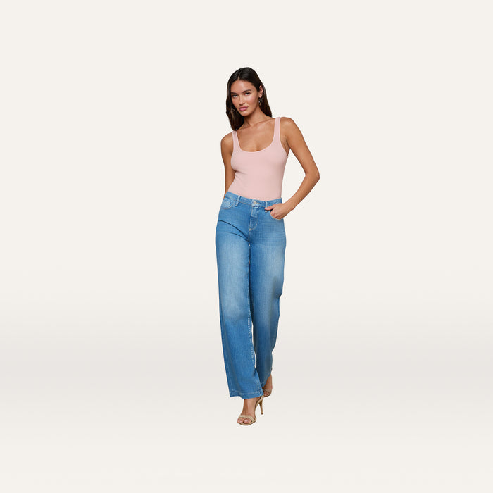 A woman poses against a plain white background wearing the Clayton Wide-Leg Jean—a high-rise, stretch denim style—in a pink tank top and heeled sandals, with one hand in her pocket.