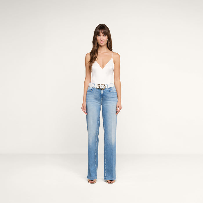 A woman stands facing forward against a plain white background, wearing the Clayton Wide-Leg Jean in light blue, paired with a white sleeveless top, a white belt with a silver buckle, and open-toe shoes.