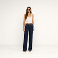 A woman wearing sunglasses, a white tank top, the Clayton Wide-Leg Jean in a dark wash, a brown belt, and brown pointed-toe shoes stands against a plain white background.