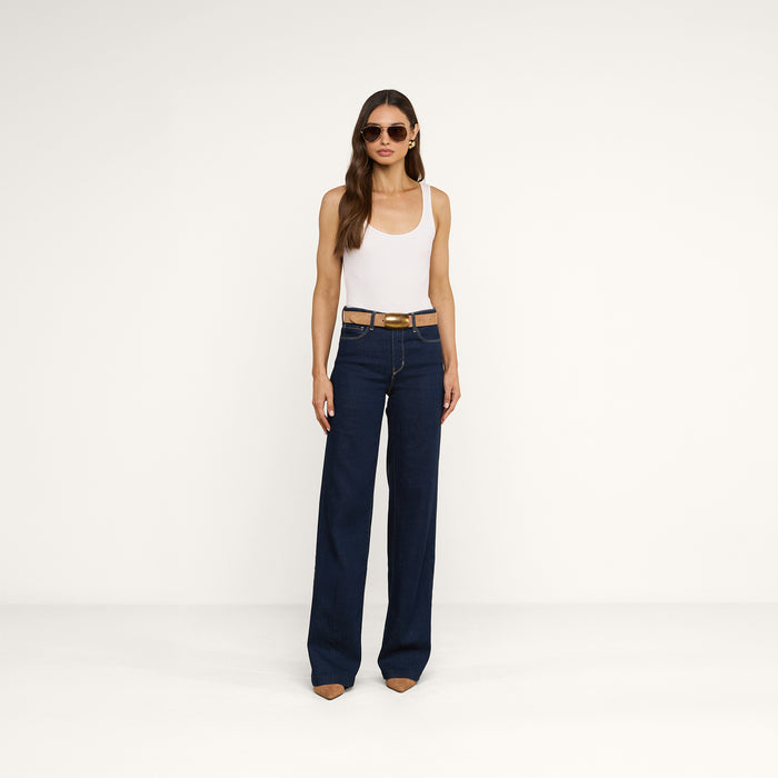 A woman wearing sunglasses, a white tank top, the Clayton Wide-Leg Jean in a dark wash, a brown belt, and brown pointed-toe shoes stands against a plain white background.