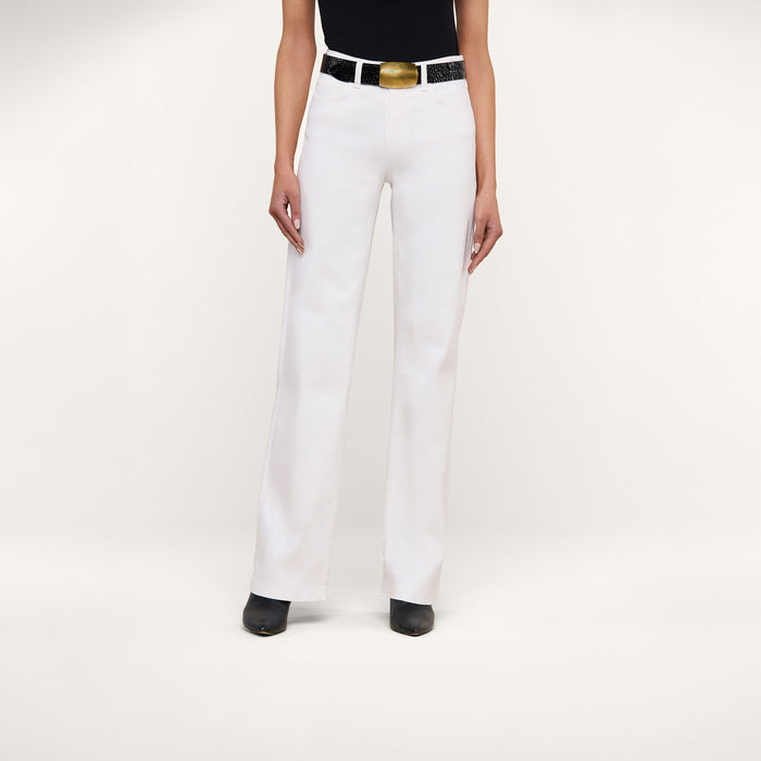 Wearing the Clayton Wide-Leg Jean in white stretch denim, paired with a black top, black boots, and a black belt featuring a large gold buckle, this person stands against a plain white background.