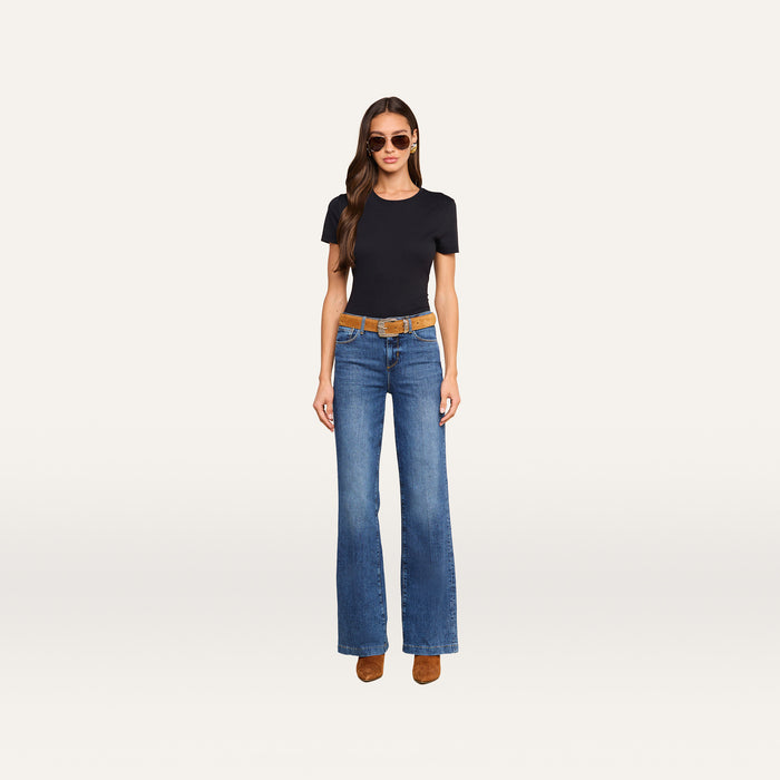 A woman wearing sunglasses, a black t-shirt, the Clayton Wide-Leg Jean in blue stretch denim, a brown belt, and brown shoes stands against a plain white background.