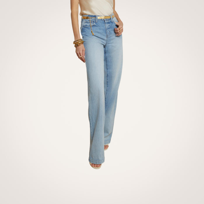 Wearing the Clayton Wide-Leg Jean in a high-rise, light wash style paired with a sleeveless beige top, gold bracelets, and a belt, this person stands against a plain, light background. Their head is not visible in the image.