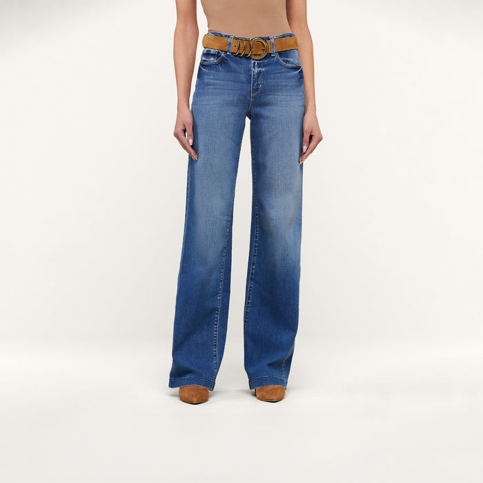 A person stands against a plain white background wearing the Clayton Wide-Leg Jean in high rise denim blue, crafted from REPREVE woven denim, styled with a brown belt and tan shoes. The upper body is cropped out of the image.