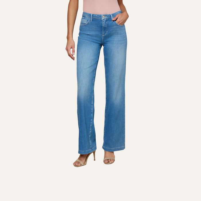 A person wearing the Clayton Wide-Leg Jean in light blue high-rise stretch denim, paired with a light pink sleeveless top and beige high-heeled sandals, stands against a plain white background.