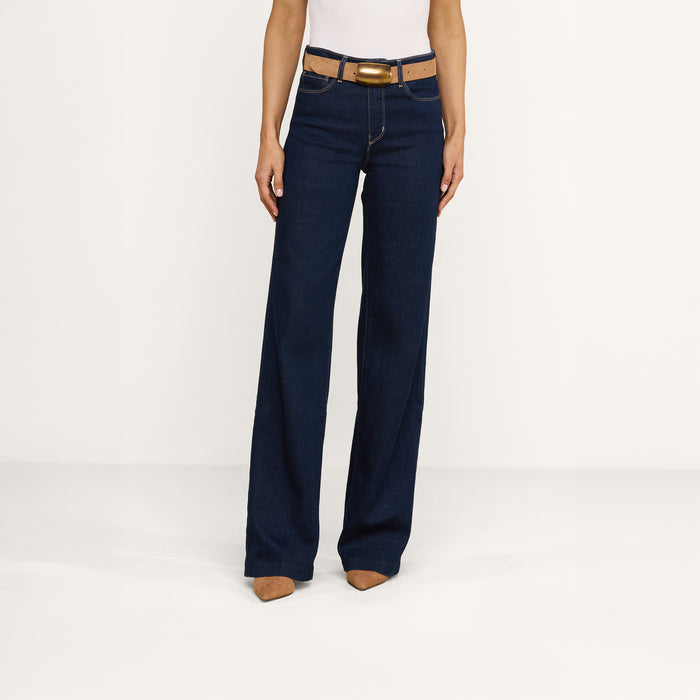 Wearing the Clayton Wide-Leg Jean, styled with a brown belt, white top, and brown pointed-toe shoes, this person is photographed from the lower torso down against a plain white background.