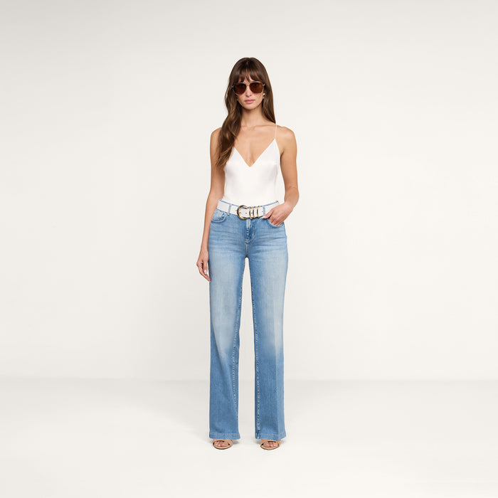 A woman wearing sunglasses, a white sleeveless top, the Clayton Wide-Leg Jean in light blue, and white sandals stands against a plain white background.