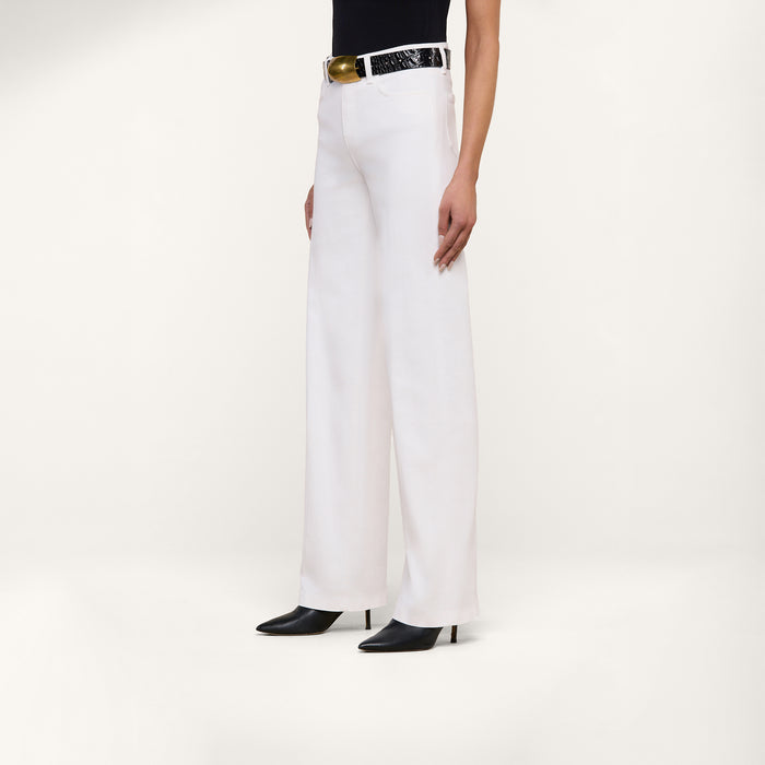 A person wearing the Clayton Wide-Leg Jean in white stretch denim, paired with a black top, a black belt with a gold buckle, and black pointed-toe shoes, stands against a plain white background.