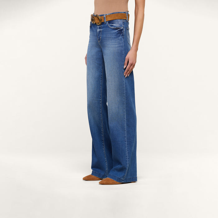 A person wearing the Clayton Wide-Leg Jean in blue high rise REPREVE woven denim, styled with a brown belt and brown pointed-toe shoes, stands against a plain white background. The upper body is mostly out of frame.