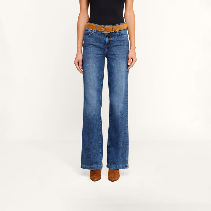A person is shown from the shoulders down wearing the Clayton Wide-Leg Jean in blue, paired with a brown belt, a black top, and brown heeled shoes, standing against a plain light background.