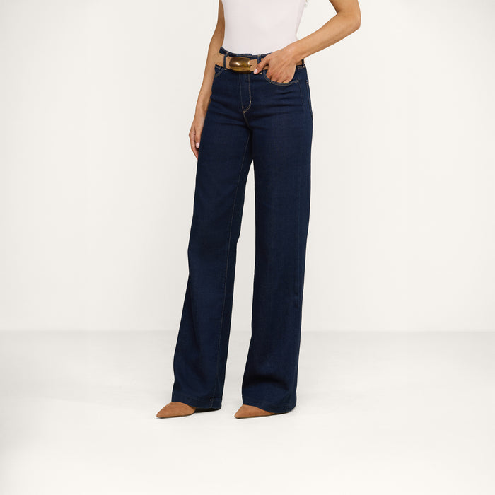 A woman stands against a plain white background wearing a sleeveless white top, the Clayton Wide-Leg Jean in a dark wash high-rise style, a brown belt, and brown pointed-toe shoes.