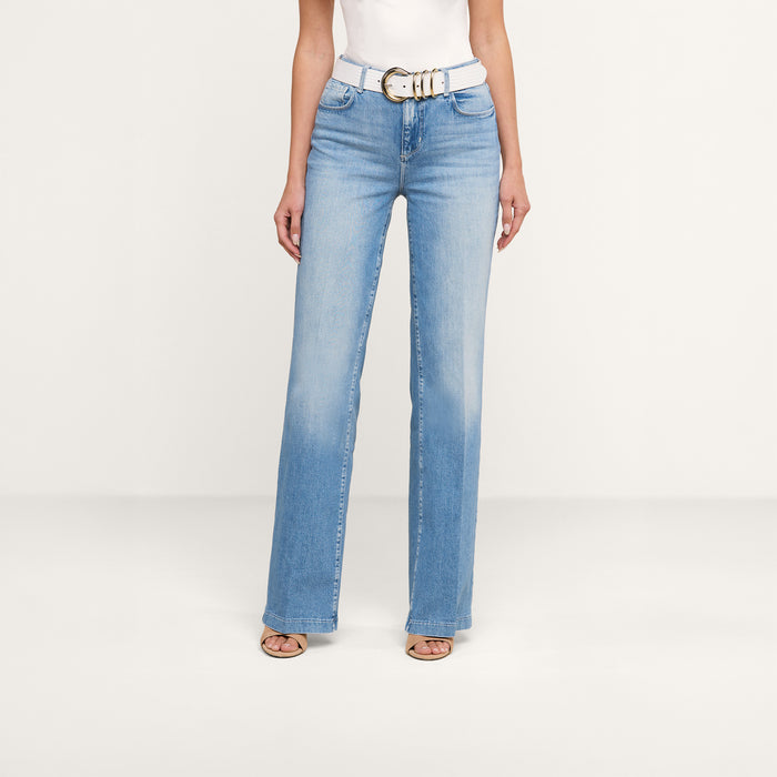 A woman wearing the Clayton Wide-Leg Jean in light blue, styled with a white belt, a white sleeveless top, and open-toe heels stands against a plain white background. Only her body from the shoulders down is visible.
