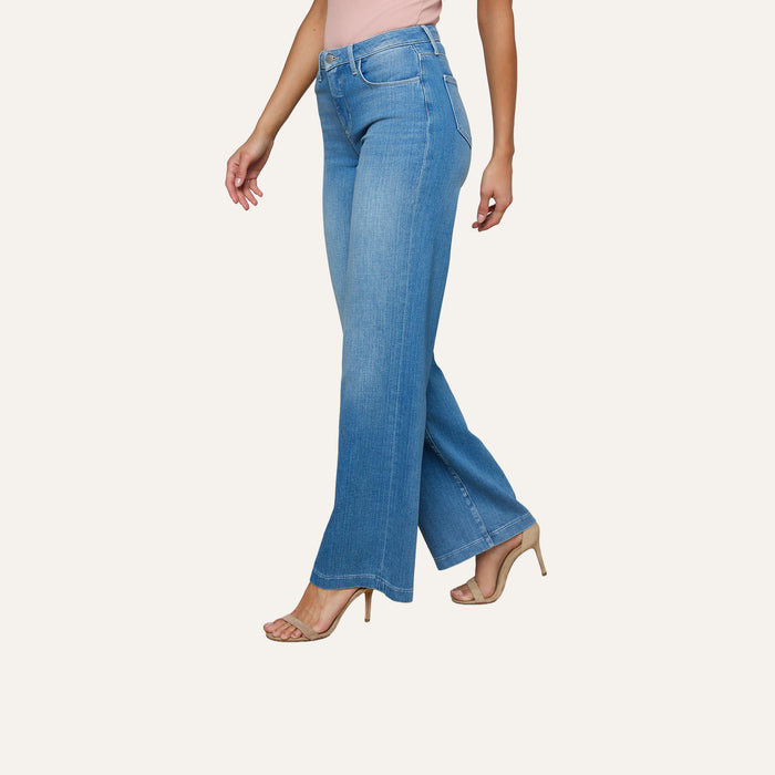 A woman is wearing the Clayton Wide-Leg Jean in light blue, featuring a high-rise and wide-leg silhouette crafted from stretch denim, along with a light pink top and nude high-heeled sandals, as she walks against a plain white background.