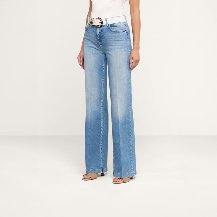 A person wearing the Clayton Wide-Leg Jean in light blue, paired with a white belt, a white sleeveless top, and open-toe heels stands against a plain white background.