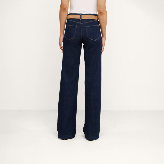 A person is standing facing away from the camera, wearing the Clayton Wide-Leg Jean in a dark wash and a white top, against a plain white background.