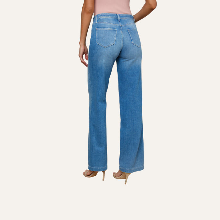 A woman is shown from the back wearing the Clayton Wide-Leg Jean in a light blue high rise style, paired with beige heels and a pink sleeveless top, her left arm bent with her hand on her hip.