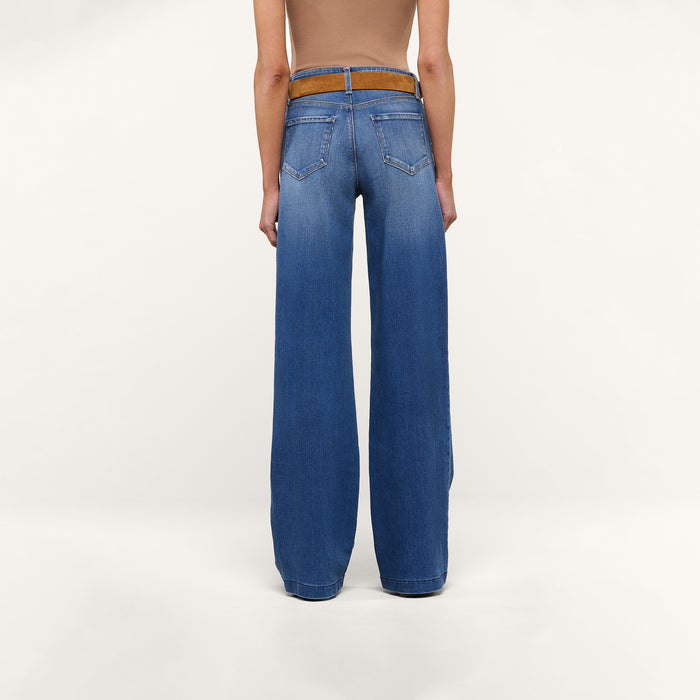A person stands facing away against a plain white background, wearing a tan sleeveless top with a brown belt and tall wide-leg blue Clayton Wide-Leg Jeans made from REPREVE woven denim.
