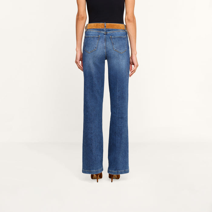 A person wearing the Clayton Wide-Leg Jean in blue, paired with a black sleeveless top, brown belt, and brown heeled shoes, is standing with their back to the camera against a plain white background.