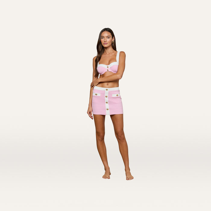 A woman stands barefoot against a plain white background, wearing the Coco Mini Skirt Cover-Up in pink bouclé with a matching textured bra top.