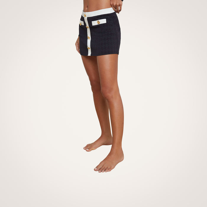 A person stands barefoot against a plain, light background, wearing the Coco Mini Skirt Cover-Up—a short, dark swim cover-up skirt with white trim and gold buttons. The upper body and head are not visible.