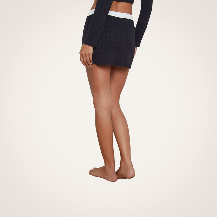 A person standing barefoot, viewed from the side and slightly from the back, wears the Coco Mini Skirt Cover-Up—a black textured bouclé fabric mini skirt with a white waistband—paired with a matching long-sleeve top. The background is plain and light.