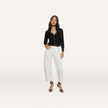 A woman stands against a plain white background, wearing the Cody Barrel-Leg Jean in high-rise white denim, paired with a black long-sleeve top and black open-toe heeled sandals. Her hair is long and loose, and she poses with one hand on her hip.
