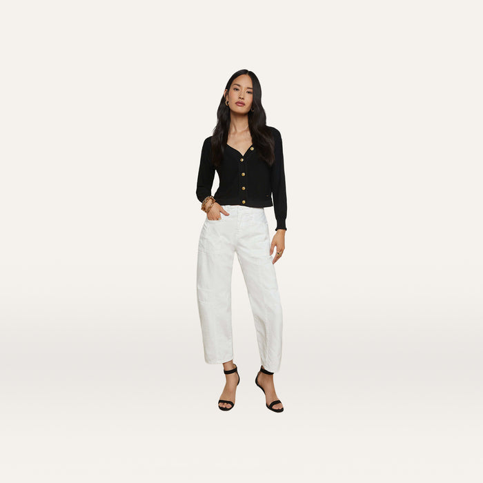 A woman stands against a plain white background, wearing the Cody Barrel-Leg Jean in high-rise white denim, paired with a black long-sleeve top and black open-toe heeled sandals. Her hair is long and loose, and she poses with one hand on her hip.
