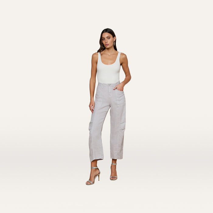A woman stands against a plain background, wearing a white tank top and the Cody Cropped Linen Pant—light gray, high-waisted, barrel-leg cargo pants made from lightweight linen and ideal for warm weather. She has long brown hair, a neutral expression, and completes the look with nude high-heeled sandals.