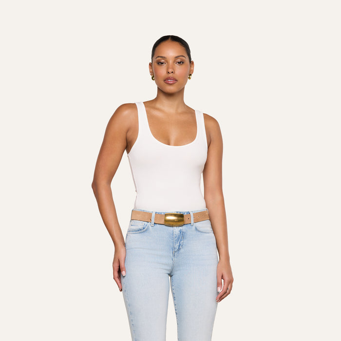A woman wearing the Cole Scoopneck Tank in white, paired with light blue jeans featuring cutouts at the waist and a gold buckle belt, stands against a plain light background.