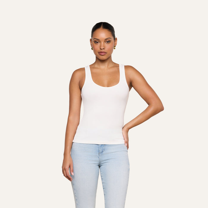 A woman stands against a plain background wearing the Cole Scoopneck Tank and light blue jeans, one arm resting on her hip and maintaining a neutral facial expression.