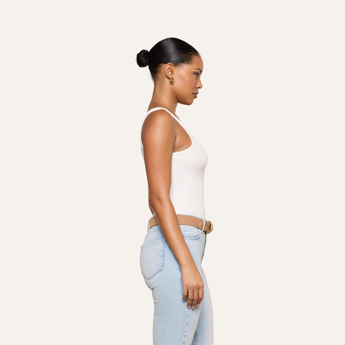 A woman wears the Cole Scoopneck Tank in white paired with light blue jeans, standing in profile against a plain, light background. Her hair is pulled back in a bun and she gazes straight ahead with a neutral expression.