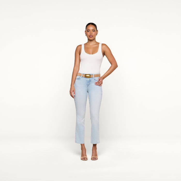 A woman stands facing forward against a plain white background, wearing the Cole Scoopneck Tank, light blue jeans, a tan belt, and nude heels. One hand rests on her hip while the other hangs by her side.