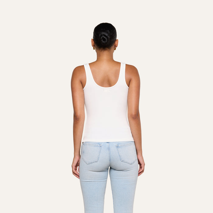 A person with dark hair in a bun wears the Cole Scoopneck Tank in white and light blue jeans, standing with their back to the camera against a plain light background.