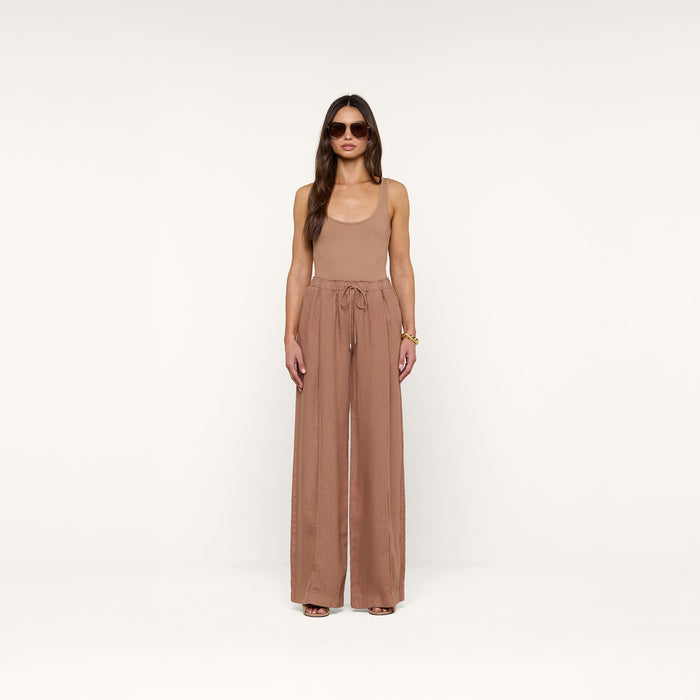 A woman stands facing forward against a plain white background, wearing sunglasses, a tan sleeveless top, and the Connie Linen Drawstring Trouser—loose, high-waisted linen pants with a drawstring waistband.