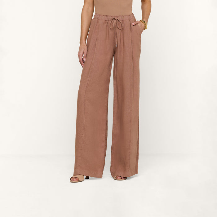 A person wearing the Connie Linen Drawstring Trouser—a wide-leg, high-waisted, light brown linen pant with a drawstring waistband—paired with a sleeveless top and heeled sandals, stands against a plain white background, with only the lower half of their body visible.